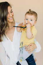 Load image into Gallery viewer, Smiling mom with baby chewing on The Collins Chewable silicone necklace in two colors tan brown wooden geometric charm black pearl beads with satin string for teething baby